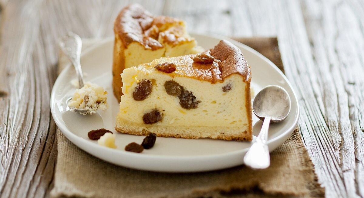 Weißer Teller, auf dem zwei Stücke Käsekuchen mit Rosinen liegen. 