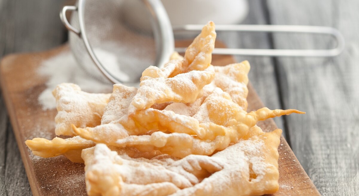 Ein Stapel frisch gebackener Faworki (auch bekannt als Engelshaar oder Krapfen), ein traditionelles polnisches Gebäck, das zu Karneval und Fasching serviert wird. Die Faworki sind goldbraun und mit Puderzucker bestäubt, der durch ein kleines Metallsieb gestreut wurde, das im Hintergrund unscharf zu sehen ist.
