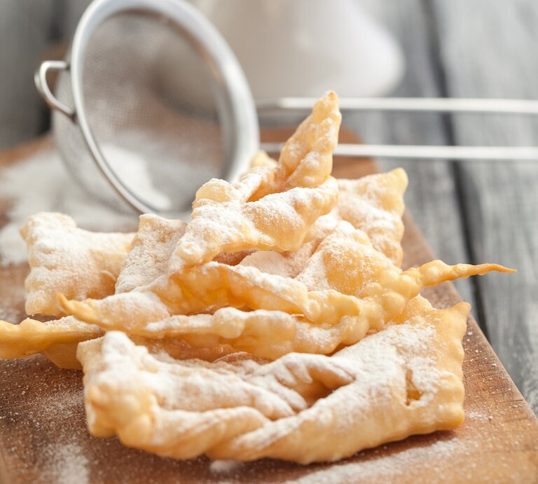 Ein Stapel frisch gebackener Faworki (auch bekannt als Engelshaar oder Krapfen), ein traditionelles polnisches Gebäck, das zu Karneval und Fasching serviert wird. Die Faworki sind goldbraun und mit Puderzucker bestäubt, der durch ein kleines Metallsieb gestreut wurde, das im Hintergrund unscharf zu sehen ist. | © iStock/ola_p