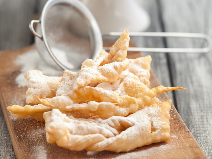 Ein Stapel frisch gebackener Faworki (auch bekannt als Engelshaar oder Krapfen), ein traditionelles polnisches Gebäck, das zu Karneval und Fasching serviert wird. Die Faworki sind goldbraun und mit Puderzucker bestäubt, der durch ein kleines Metallsieb gestreut wurde, das im Hintergrund unscharf zu sehen ist. | © iStock/ola_p