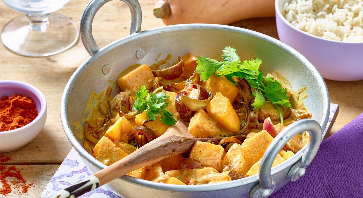Kürbis-Curry in einer weißen Pfanne auf hölzernem Hintergrund. Rechts eine Schüssel mit Reis, links Gewürze und ein Wasserglas. Oben auf dem Foto ein Kürbis.