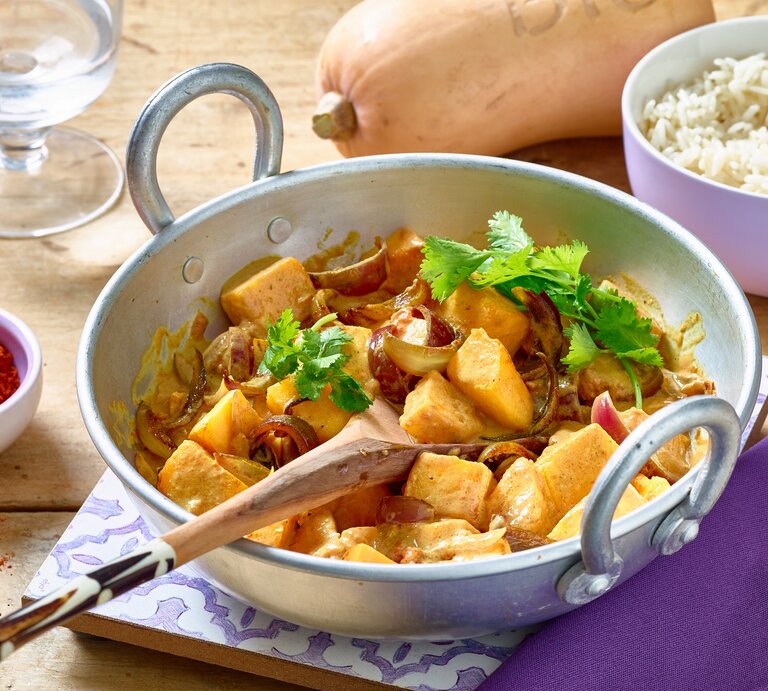 Kürbis-Curry in einer weißen Pfanne auf hölzernem Hintergrund. Rechts eine Schüssel mit Reis, links Gewürze und ein Wasserglas. Oben auf dem Foto ein Kürbis. | © Matthias Liebich