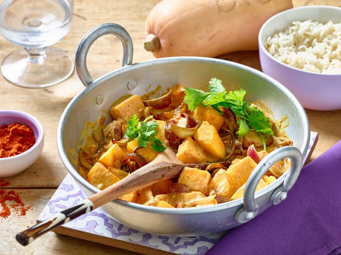 Kürbis-Curry in einer weißen Pfanne auf hölzernem Hintergrund. Rechts eine Schüssel mit Reis, links Gewürze und ein Wasserglas. Oben auf dem Foto ein Kürbis. | © Matthias Liebich
