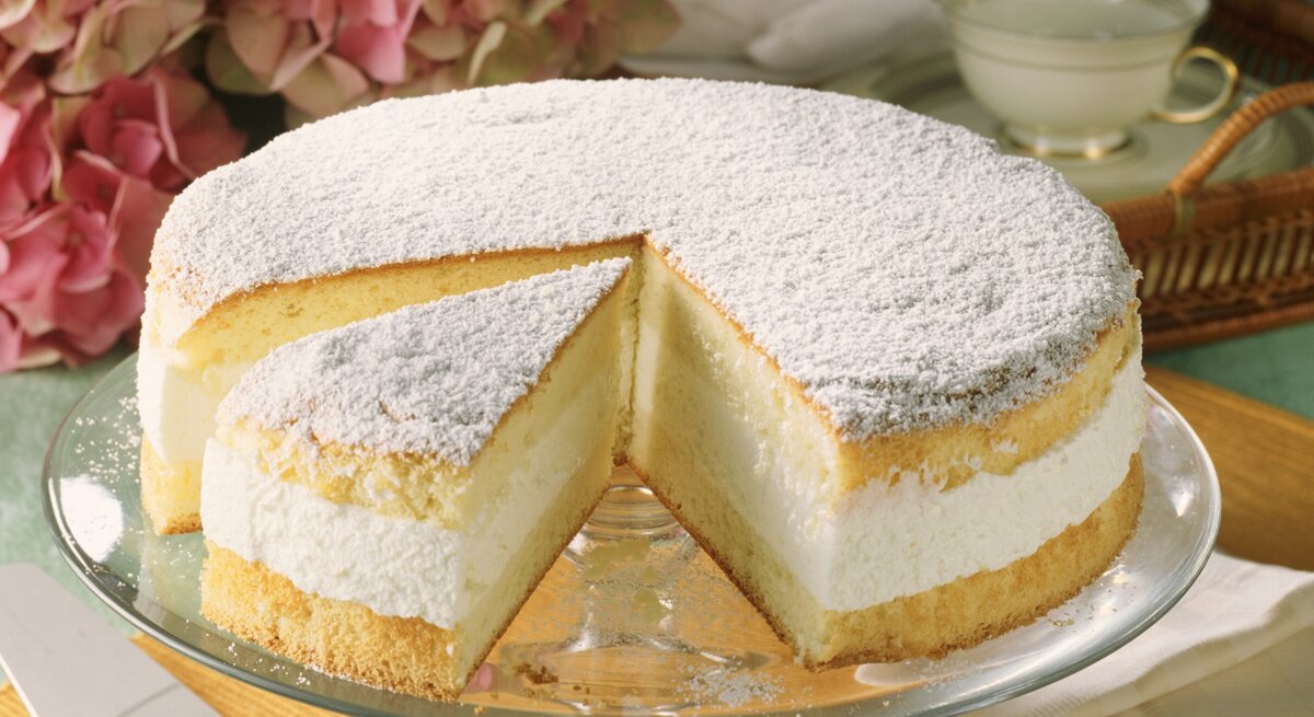 Die Käse-Sahne-Torte auf einem grünlichen Tisch mit pinken Blüten im Hintergrund.