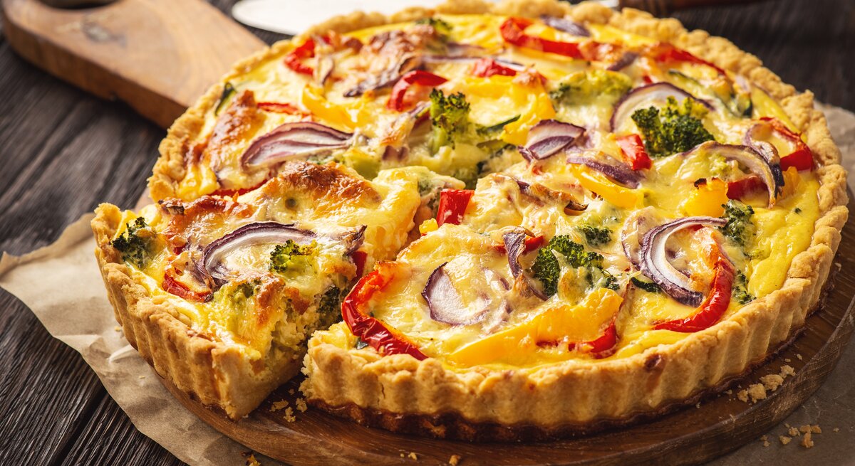 Das ist ein Bild von einer herzhaften Torte, oft als Quiche bekannt. Es sieht aus, als wäre es mit einer Vielzahl von Gemüse gefüllt, darunter Brokkoli, rote Paprika und rote Zwiebeln. Die Torte hat eine goldbraune Kruste und das Füllung ist gut gebacken, wobei einige Teile des Gemüses leicht gebräunt sind, was auf eine knusprige Textur hinweist. Die Farben sind lebhaft und machen das Gericht optisch ansprechend.