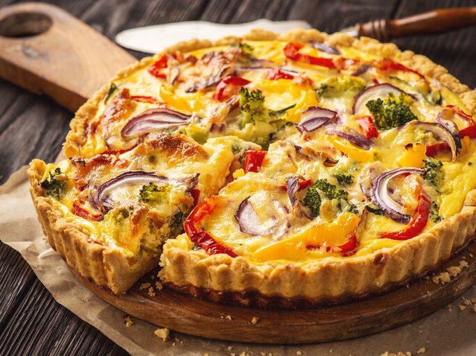 Das ist ein Bild von einer herzhaften Torte, oft als Quiche bekannt. Es sieht aus, als wäre es mit einer Vielzahl von Gemüse gefüllt, darunter Brokkoli, rote Paprika und rote Zwiebeln. Die Torte hat eine goldbraune Kruste und das Füllung ist gut gebacken, wobei einige Teile des Gemüses leicht gebräunt sind, was auf eine knusprige Textur hinweist. Die Farben sind lebhaft und machen das Gericht optisch ansprechend. | © iStock.com/iko636