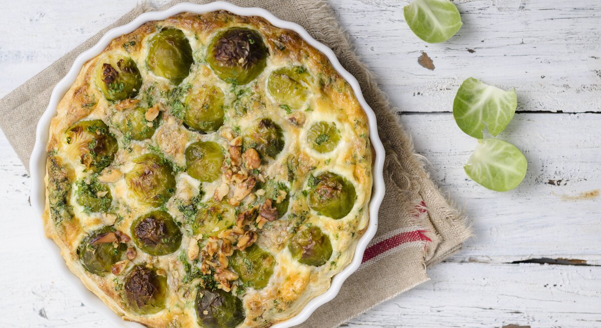 Draufsicht auf eine weiße Tarteform gefüllt mit Rosenkohlquiche. Das Ganze steht auf porösem, weißen Holz. 