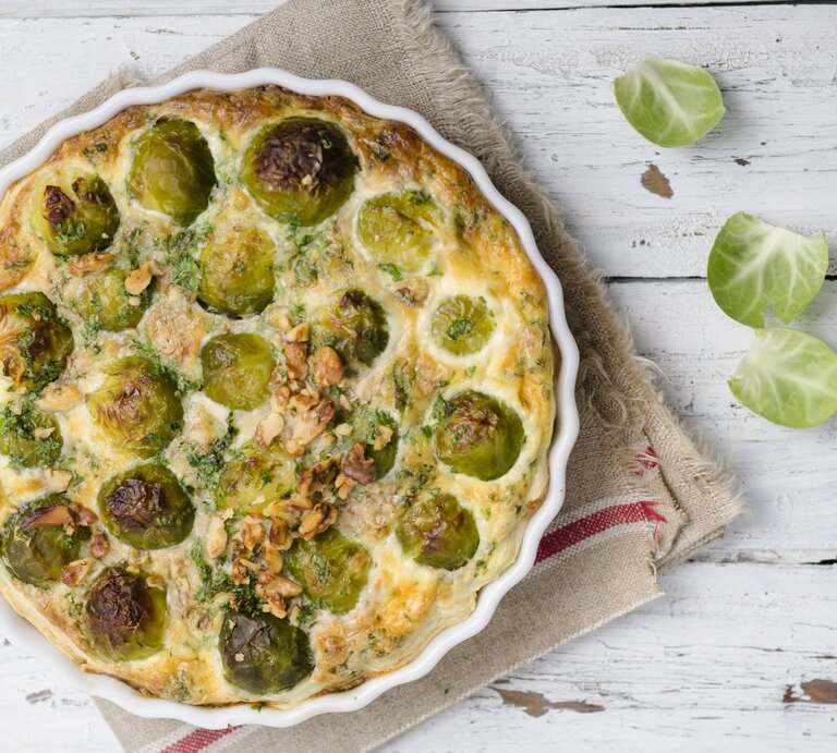 Draufsicht auf eine weiße Tarteform gefüllt mit Rosenkohlquiche. Das Ganze steht auf porösem, weißen Holz.  | © imago images/Westend61