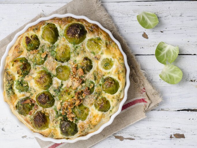 Draufsicht auf eine weiße Tarteform gefüllt mit Rosenkohlquiche. Das Ganze steht auf porösem, weißen Holz.  | © imago images/Westend61