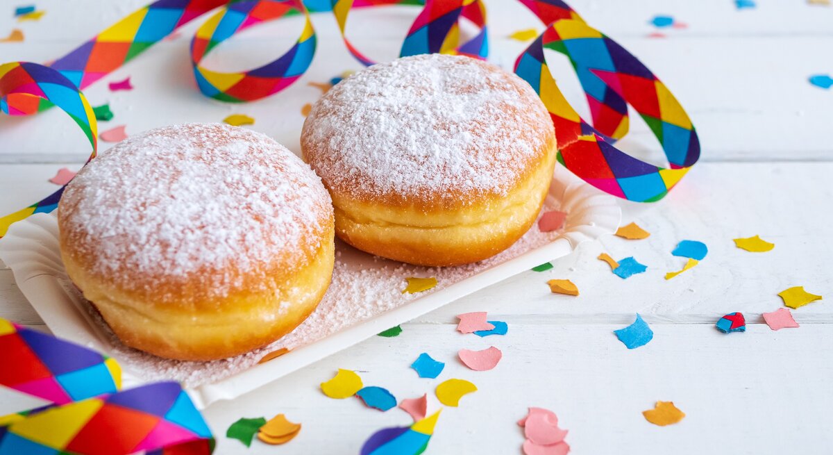 zwei Berliner mit Puderzucker bestäubt in weißem Szenario. Daneben Konfetti und Luftschlangen.