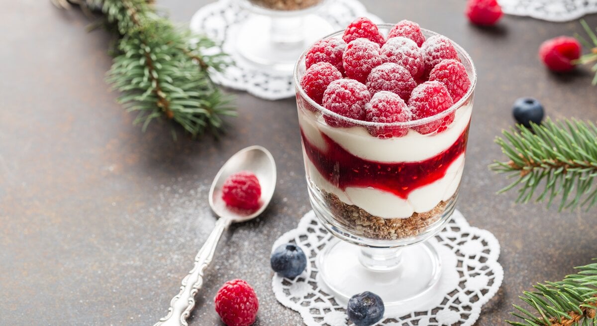 Dessert im Glas mit Himbeeren und Creme mit Kuchenboden auf dunklem Untergrund. Umgeben von Tannenzweigen.