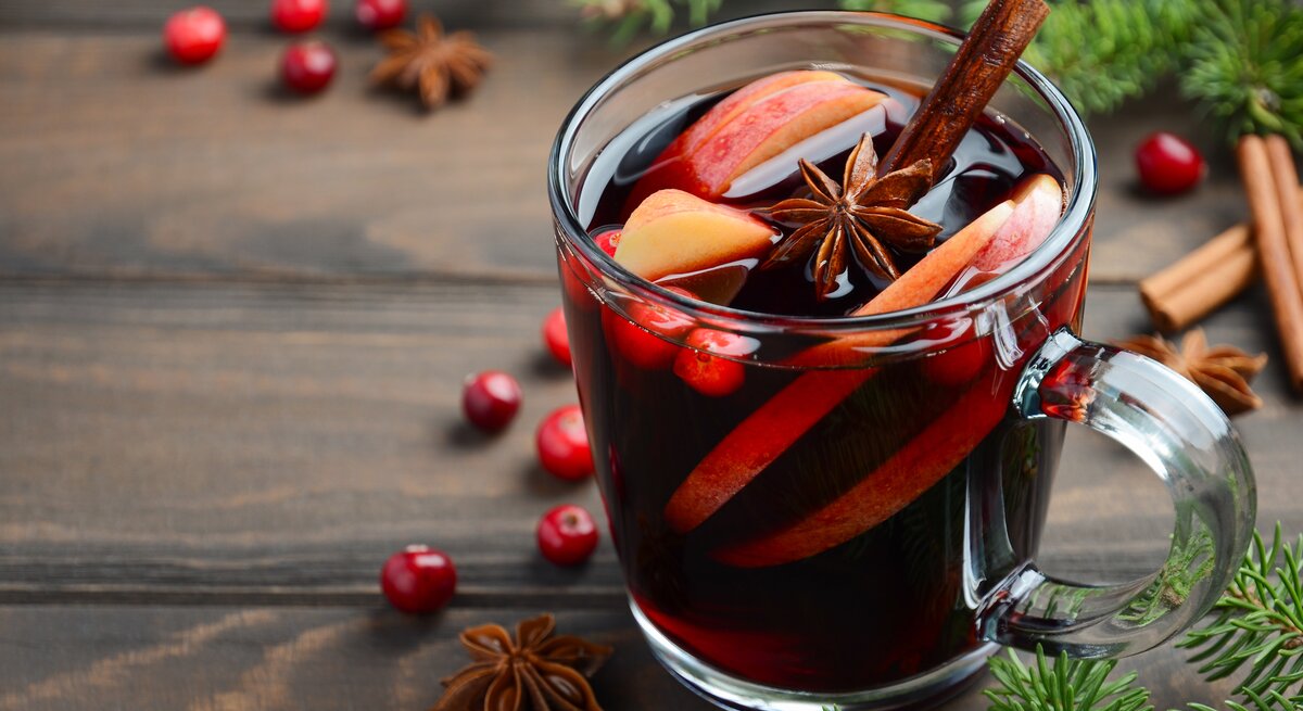 Eine durchsichtige Tasse gefüllt mit selbst gemachtem Glühwein auf Holztisch. Daneben weihnachtliche Gewürze und Deko-Elemente. 