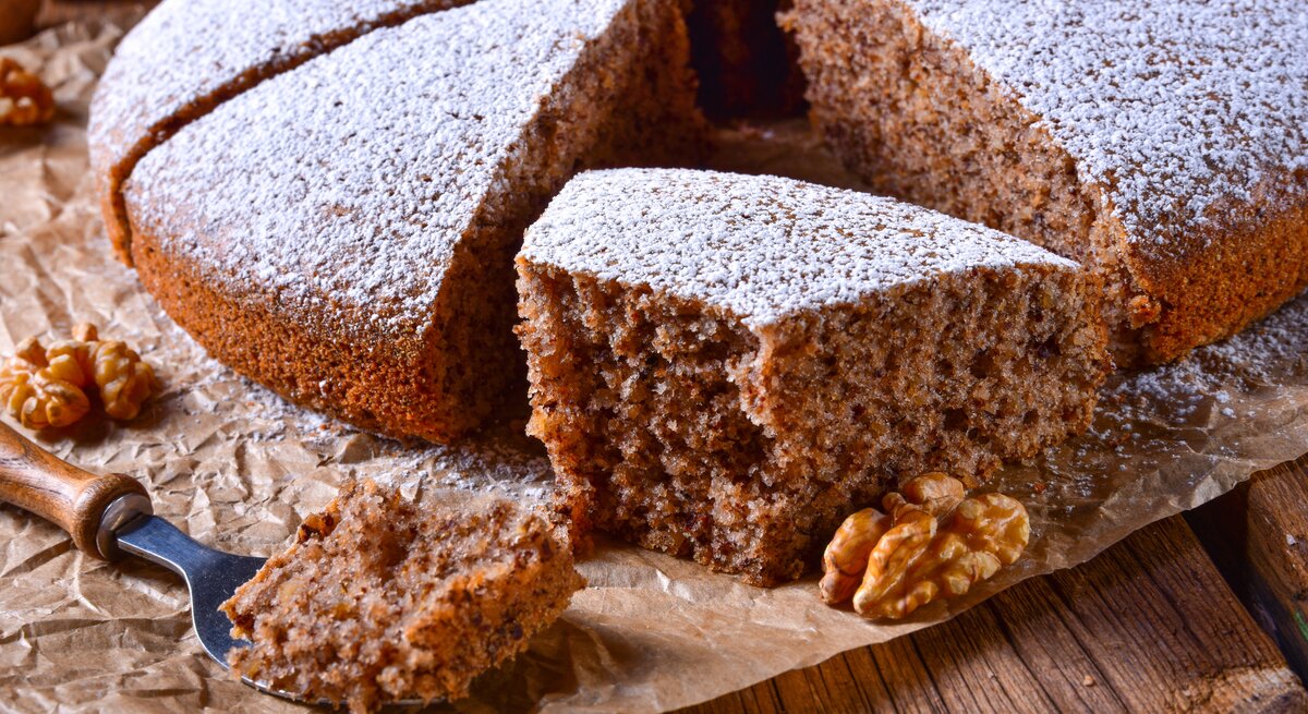 Ein angeschnittener Walnusskuchen mit Schokolade, bestäubt mit Puderzucker, auf einem Holztisch mit Backpapier. 