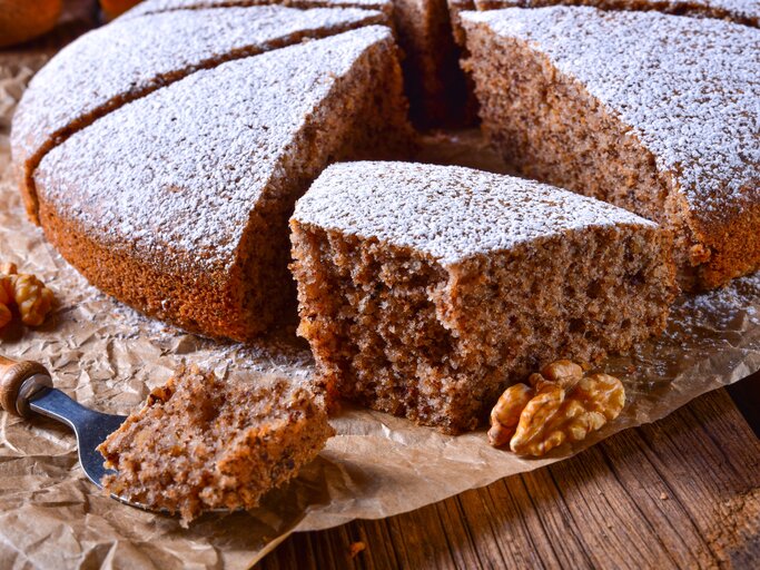 Ein angeschnittener Walnusskuchen mit Schokolade, bestäubt mit Puderzucker, auf einem Holztisch mit Backpapier.  | © iStock.com/Dar1930