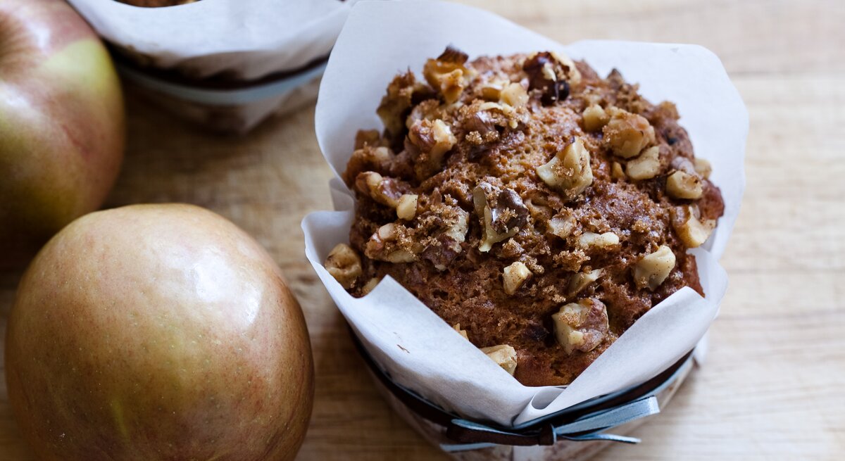 Ein saftiger Apfel-Walnuss-Muffin in weißem Papier liegt auf einem Holztisch, daneben zwei frische, rote Äpfel.