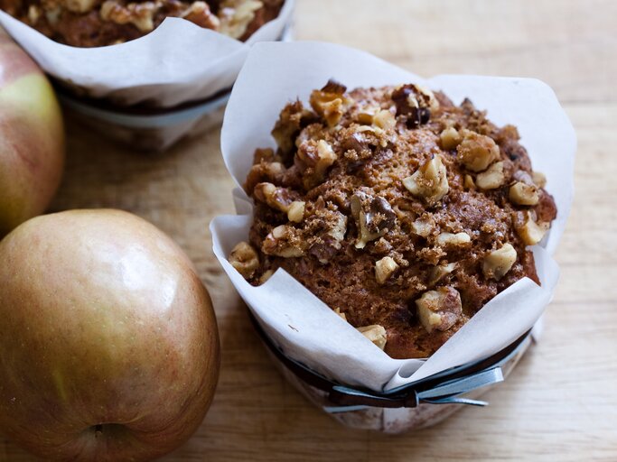Ein saftiger Apfel-Walnuss-Muffin in weißem Papier liegt auf einem Holztisch, daneben zwei frische, rote Äpfel. | © Getty Images/KitchenM