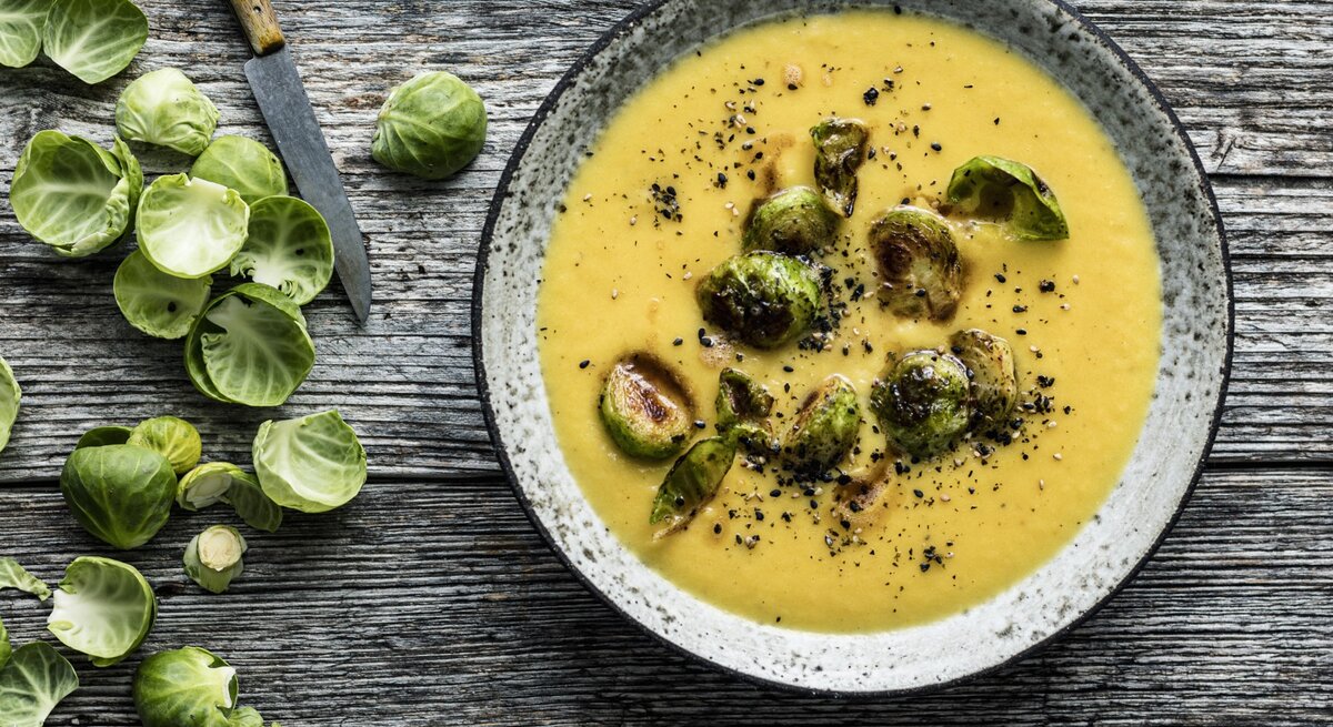 Draufsicht: Hölzerner Untergrund auf der ein grauer Steingutteller mit Steckrübensuppe und Rosenkohl-Topping steht. Links daneben noch Schalen frischer Rosenkohlröschen und ein scharfes Messer mit Holzgriff.