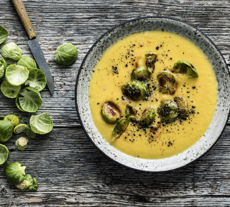Draufsicht: Hölzerner Untergrund auf der ein grauer Steingutteller mit Steckrübensuppe und Rosenkohl-Topping steht. Links daneben noch Schalen frischer Rosenkohlröschen und ein scharfes Messer mit Holzgriff. | © StockFood / Gräfe & Unzer Verlag / Schardt, Wolfgang