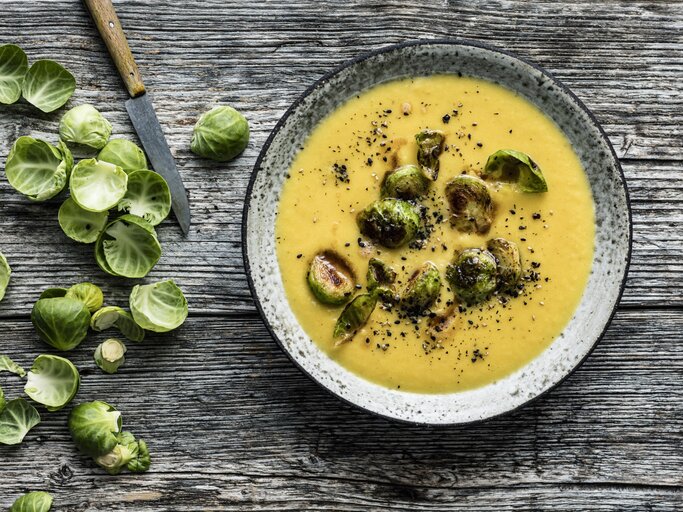 Draufsicht: Hölzerner Untergrund auf der ein grauer Steingutteller mit Steckrübensuppe und Rosenkohl-Topping steht. Links daneben noch Schalen frischer Rosenkohlröschen und ein scharfes Messer mit Holzgriff. | © StockFood / Gräfe & Unzer Verlag / Schardt, Wolfgang