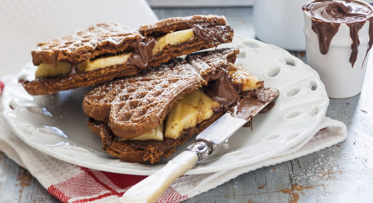 Schoko-Waffeln mit Banane auf einem weißen Teller auf einem Geschirrtuch. Auf dem Teller liegt ein Messer und hinten auf dem Bild ein Glas mit Schoko-Creme.