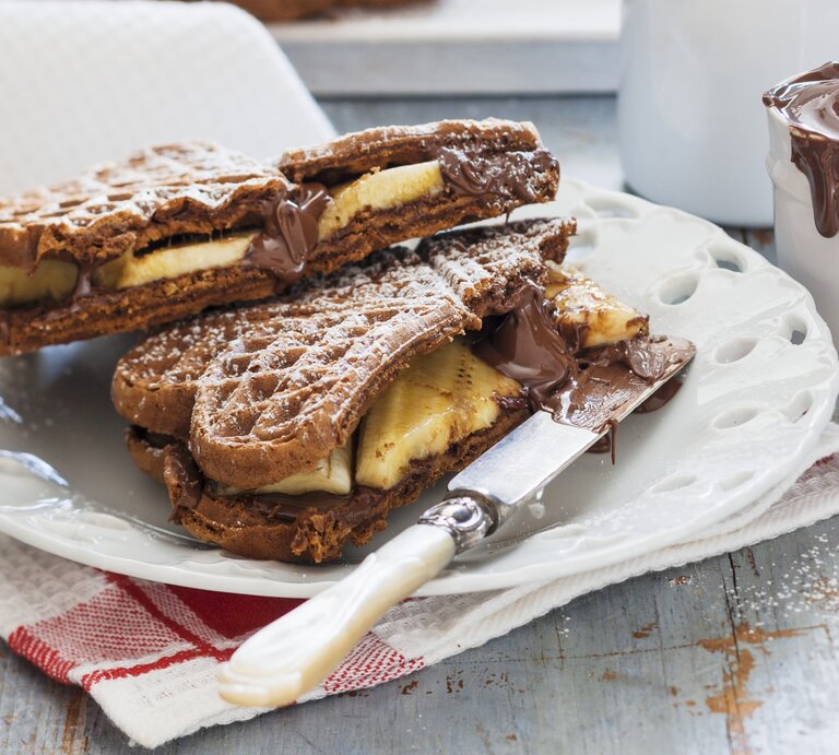 Schoko-Waffeln mit Banane auf einem weißen Teller auf einem Geschirrtuch. Auf dem Teller liegt ein Messer und hinten auf dem Bild ein Glas mit Schoko-Creme. | © StockFood / Young, Andrew
