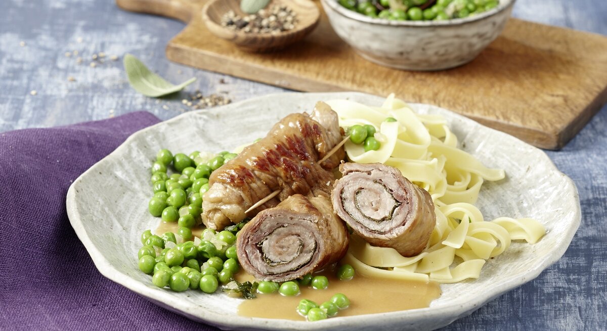 Ein Teller Saltimbocca Kalbsrouladen, dazu breite Bandnudeln und grüne Erbsen, angerichtet auf einer hellen Keramikplatte.