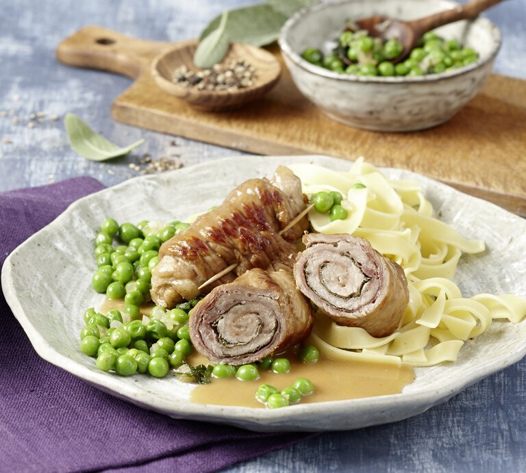 Ein Teller Saltimbocca Kalbsrouladen, dazu breite Bandnudeln und grüne Erbsen, angerichtet auf einer hellen Keramikplatte. | © EAT CLUB/Isabell Triemer