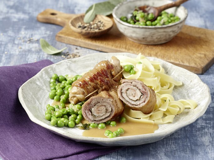 Ein Teller Saltimbocca Kalbsrouladen, dazu breite Bandnudeln und grüne Erbsen, angerichtet auf einer hellen Keramikplatte. | © EAT CLUB/Isabell Triemer