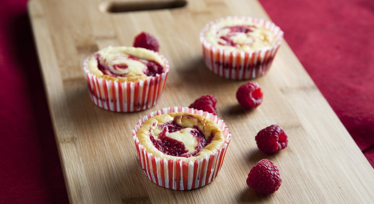 Drei Käsekuchen-Muffins mit Himbeeren arrangiert auf einem Holzbrett. 
