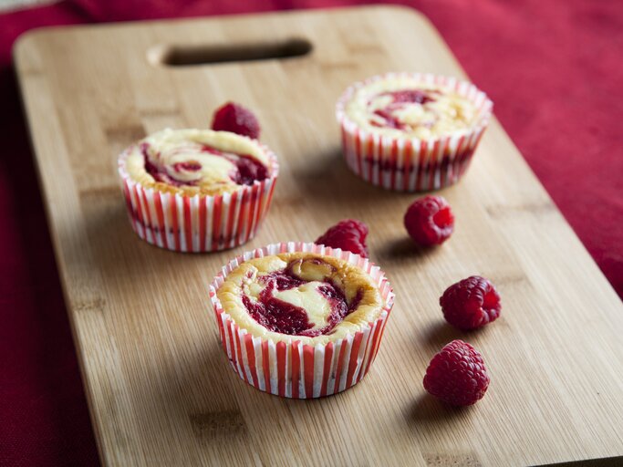 Drei Käsekuchen-Muffins mit Himbeeren arrangiert auf einem Holzbrett.  | © iStock/cheche22