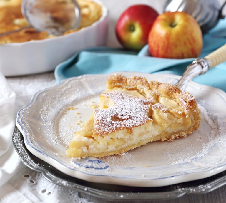 Ein Stück Apfelkuchen mit Puderzucker bestäubt liegt auf einem Teller, im Hintergrund ein ganzer Kuchen und zwei Äpfel. | © iStock.com/photosimysia