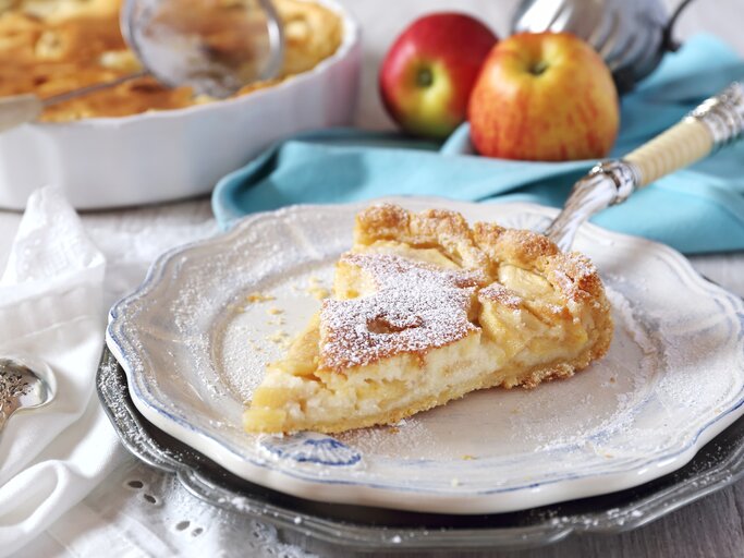 Ein Stück Apfelkuchen mit Puderzucker bestäubt liegt auf einem Teller, im Hintergrund ein ganzer Kuchen und zwei Äpfel. | © iStock.com/photosimysia