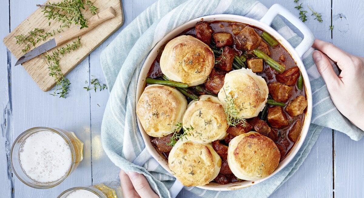 Bier-Gulasch mit Brötchen in einem weißen Topf auf hellem Hintergrund.