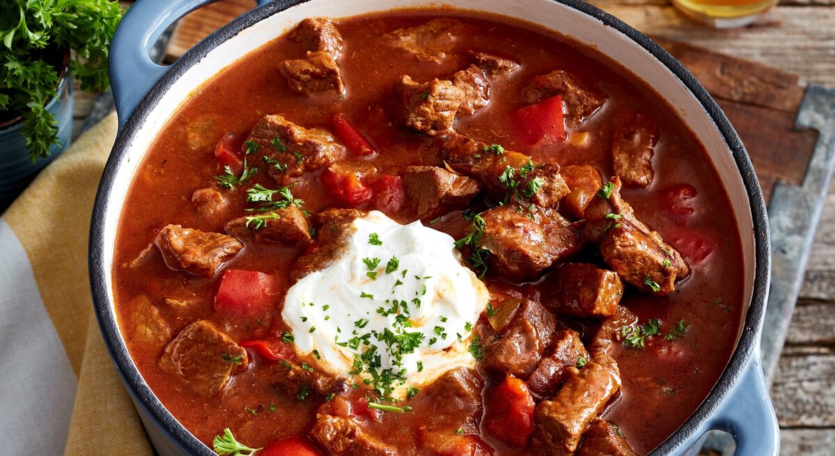Brauhaus-Gulasch mit Kartoffel-Pastinaken-Stampf und Schmand in einem Topf mit blauen Griffen auf Holzbrett und Holztisch. Im Bild ein Glas mit Petersilie und ein Glas Bier angedeutet. Draufsicht.
