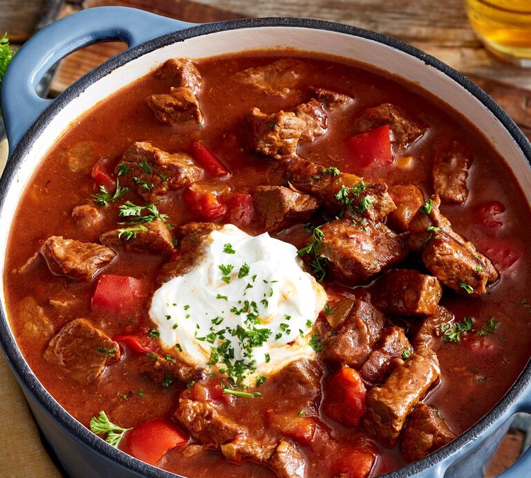 Brauhaus-Gulasch mit Kartoffel-Pastinaken-Stampf und Schmand in einem Topf mit blauen Griffen auf Holzbrett und Holztisch. Im Bild ein Glas mit Petersilie und ein Glas Bier angedeutet. Draufsicht. | © EAT CLUB/Isabell Triemer