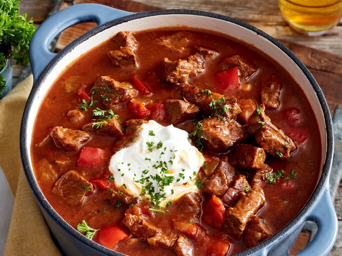 Brauhaus-Gulasch mit Kartoffel-Pastinaken-Stampf und Schmand in einem Topf mit blauen Griffen auf Holzbrett und Holztisch. Im Bild ein Glas mit Petersilie und ein Glas Bier angedeutet. Draufsicht. | © EAT CLUB/Isabell Triemer