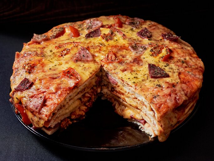 Gestapelter Pizzakuchen mit Käse, Schinken und Salami in dunkler Kulisse.  | © GettyImages/Maya Malkova