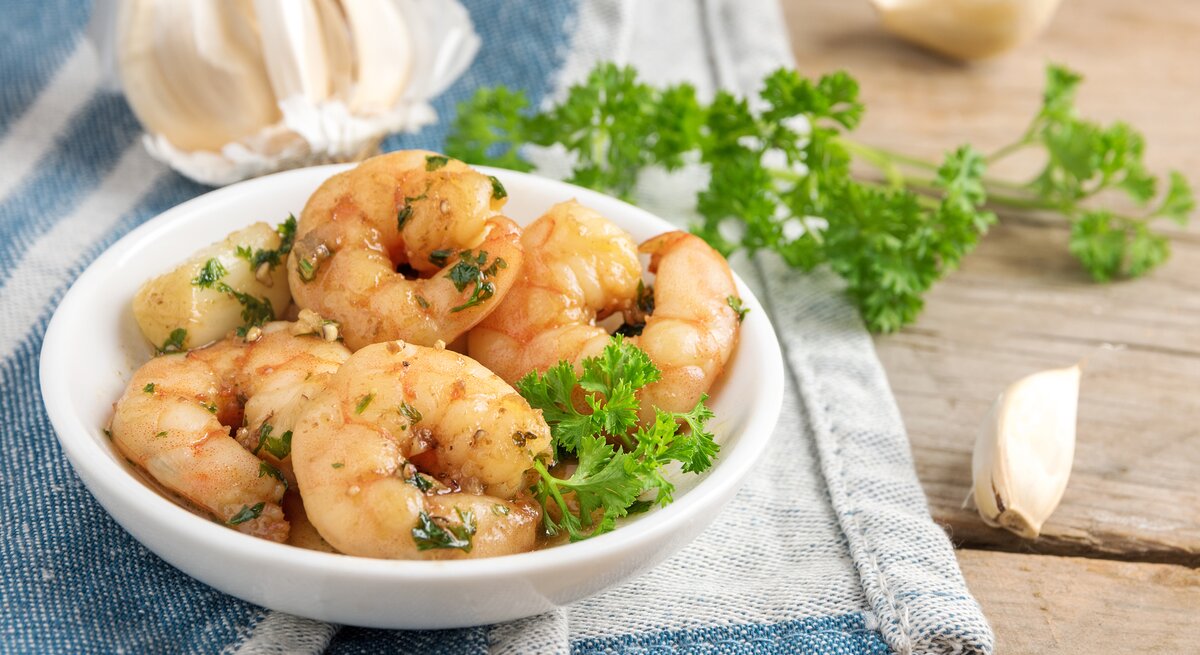 Mehrere Gambas von Ajo liegen in einer weißen Schüssel auf einem Geschirrhandtuch auf einem Holztisch. Um die Schüssel herum liegen Knoblauch und Petersilie.