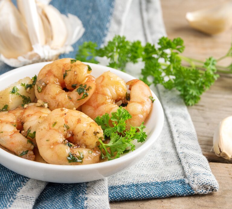 Mehrere Gambas von Ajo liegen in einer weißen Schüssel auf einem Geschirrhandtuch auf einem Holztisch. Um die Schüssel herum liegen Knoblauch und Petersilie. | © Getty Images / Maren Winter / EyeEm