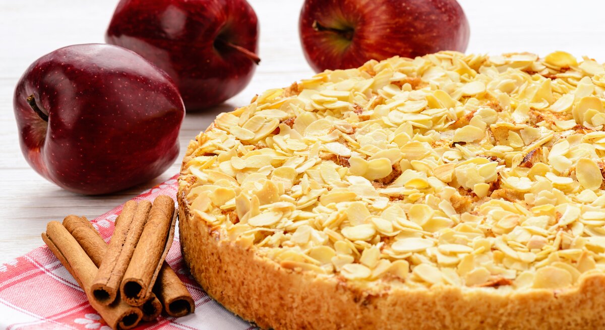 Apfelkuchen mit Mandeln vor weißem Hintergrund. Daneben liegen Äpfel und Zimtstangen. 