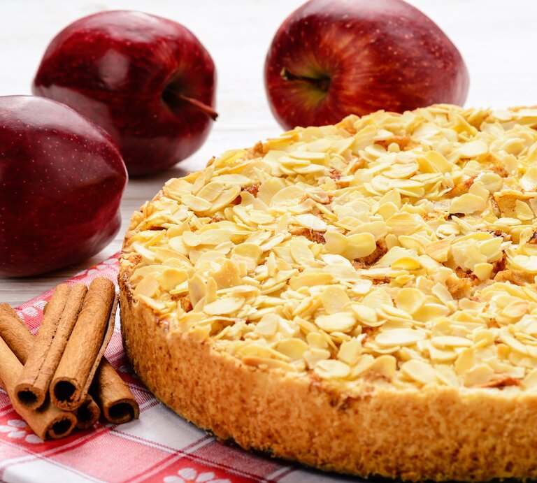 Apfelkuchen mit Mandeln vor weißem Hintergrund. Daneben liegen Äpfel und Zimtstangen.  | © iStock.com/iko636