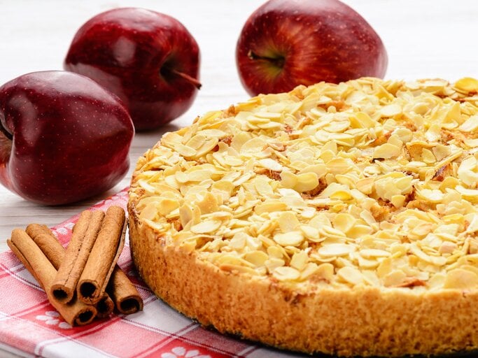 Apfelkuchen mit Mandeln vor weißem Hintergrund. Daneben liegen Äpfel und Zimtstangen.  | © iStock.com/iko636