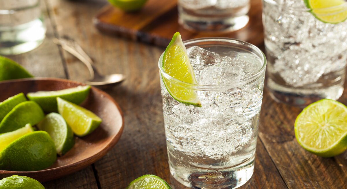Glas mit prickelnder, durchsichtiger Flüssigkeit (Gin Tonic) dekortiert mit Limette auf Holztisch