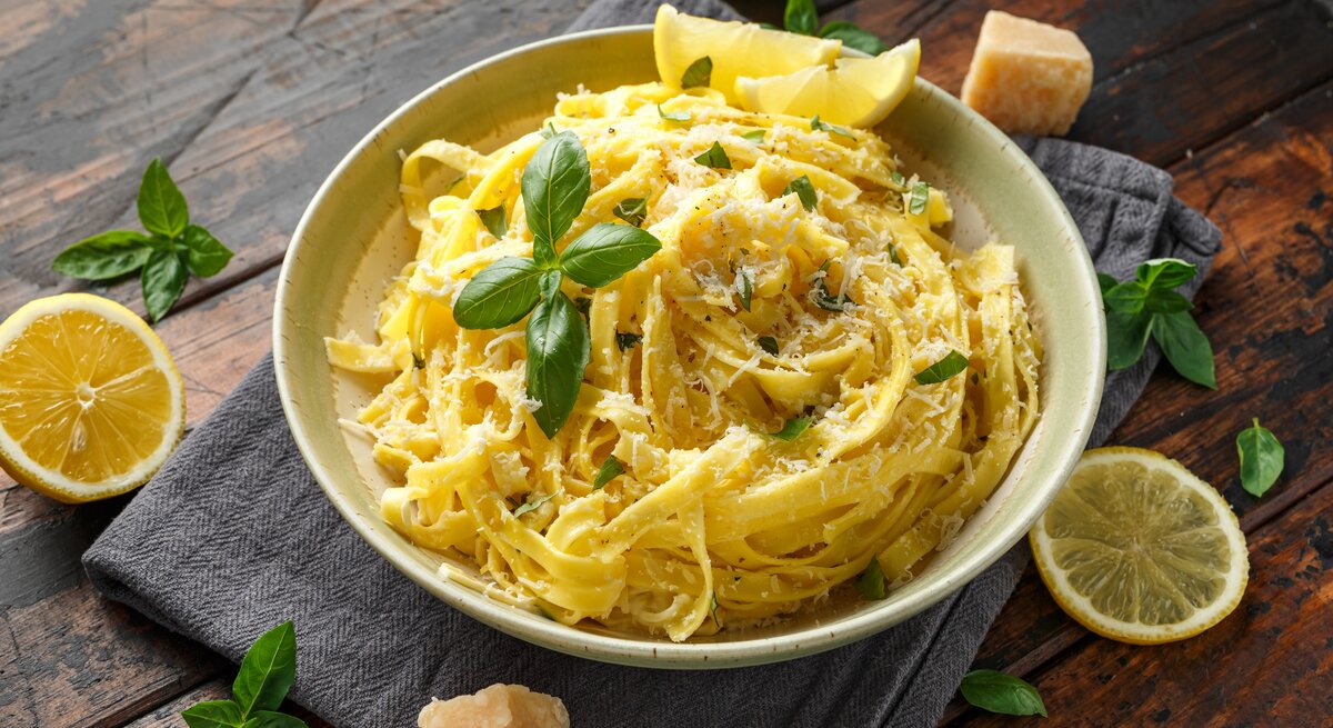 Ein Teller mit Linguine al Limone in einer cremigen Zitronensauce, garniert mit frisch geriebenem Parmesan und Basilikumblättern. Neben dem Teller liegen Zitronenscheiben, frische Basilikumblätter und ein Stück Parmesan auf einem dunklen Holztisch.