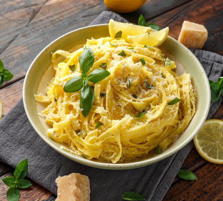 Ein Teller mit Linguine al Limone in einer cremigen Zitronensauce, garniert mit frisch geriebenem Parmesan und Basilikumblättern. Neben dem Teller liegen Zitronenscheiben, frische Basilikumblätter und ein Stück Parmesan auf einem dunklen Holztisch. | © GettyImages.de/DronG