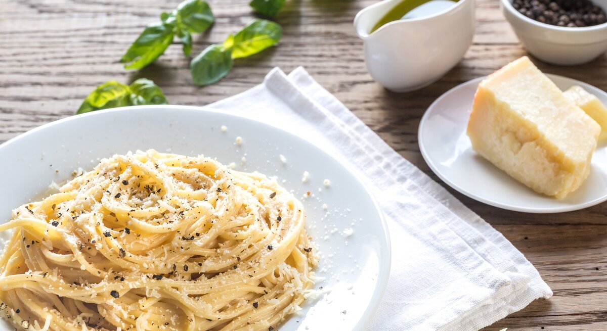 Ein Teller Pasta Cacio e Pepe auf einem rustikalen Holztisch mit Zutaten wie Pfeffer und Parmesan. Alles von schräg oben fotografiert.