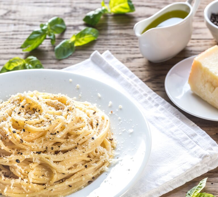 Ein Teller Pasta Cacio e Pepe auf einem rustikalen Holztisch mit Zutaten wie Pfeffer und Parmesan. Alles von schräg oben fotografiert. | © gettyimages.de / AlexPro9500