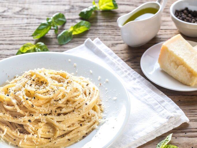 Ein Teller Pasta Cacio e Pepe auf einem rustikalen Holztisch mit Zutaten wie Pfeffer und Parmesan. Alles von schräg oben fotografiert. | © gettyimages.de / AlexPro9500