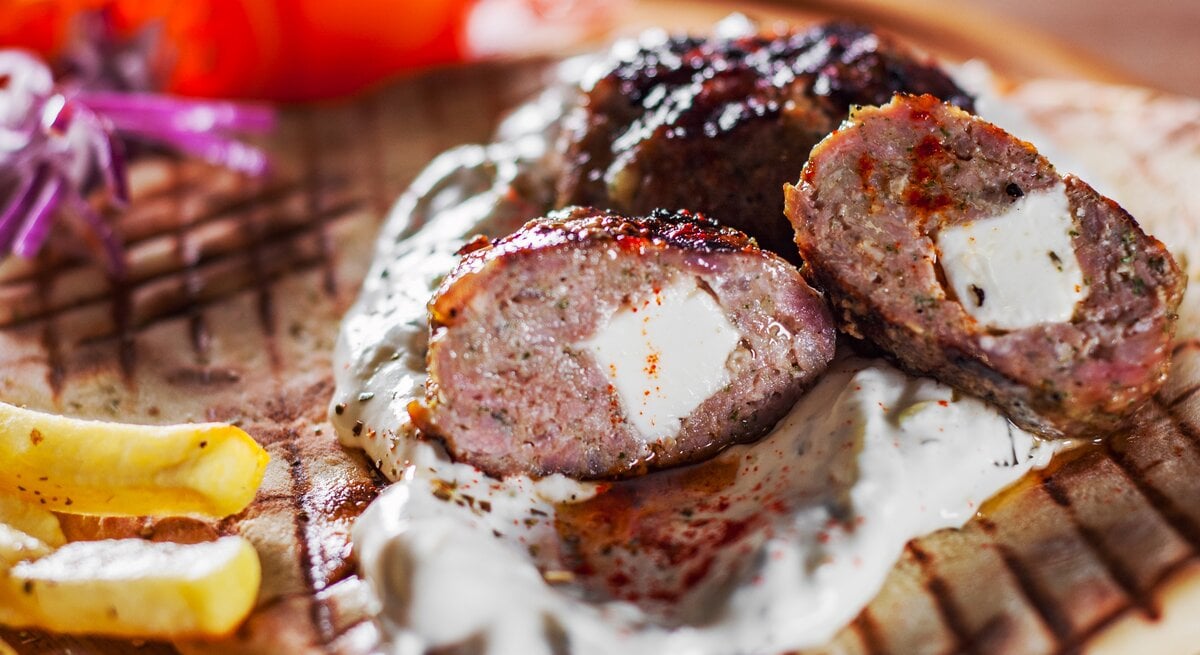 Angeschnittenes, griechisches Hacksteak "Bifteki" mit Feta-Füllung. Darunter ein Fladenbrot, daneben Pommes Frites. 