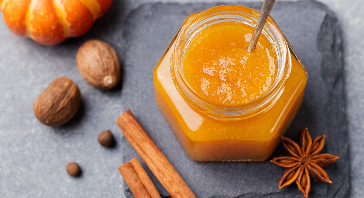 Ein Glas mit Kürbis-Orangen-Marmelade steht auf einer Steinplatte auf einem grauen Untergrund. Neben dem Glas liegt Anis und zwei Zimtstangen. Neben der Steinplatten liegen einige Nüsse und ein Zierkürbis. 